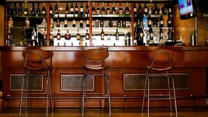 Un espace bar avec des panneaux de bois, de nombreuses étagères remplies de bouteilles d’alcool et trois tabourets de bar en métal.