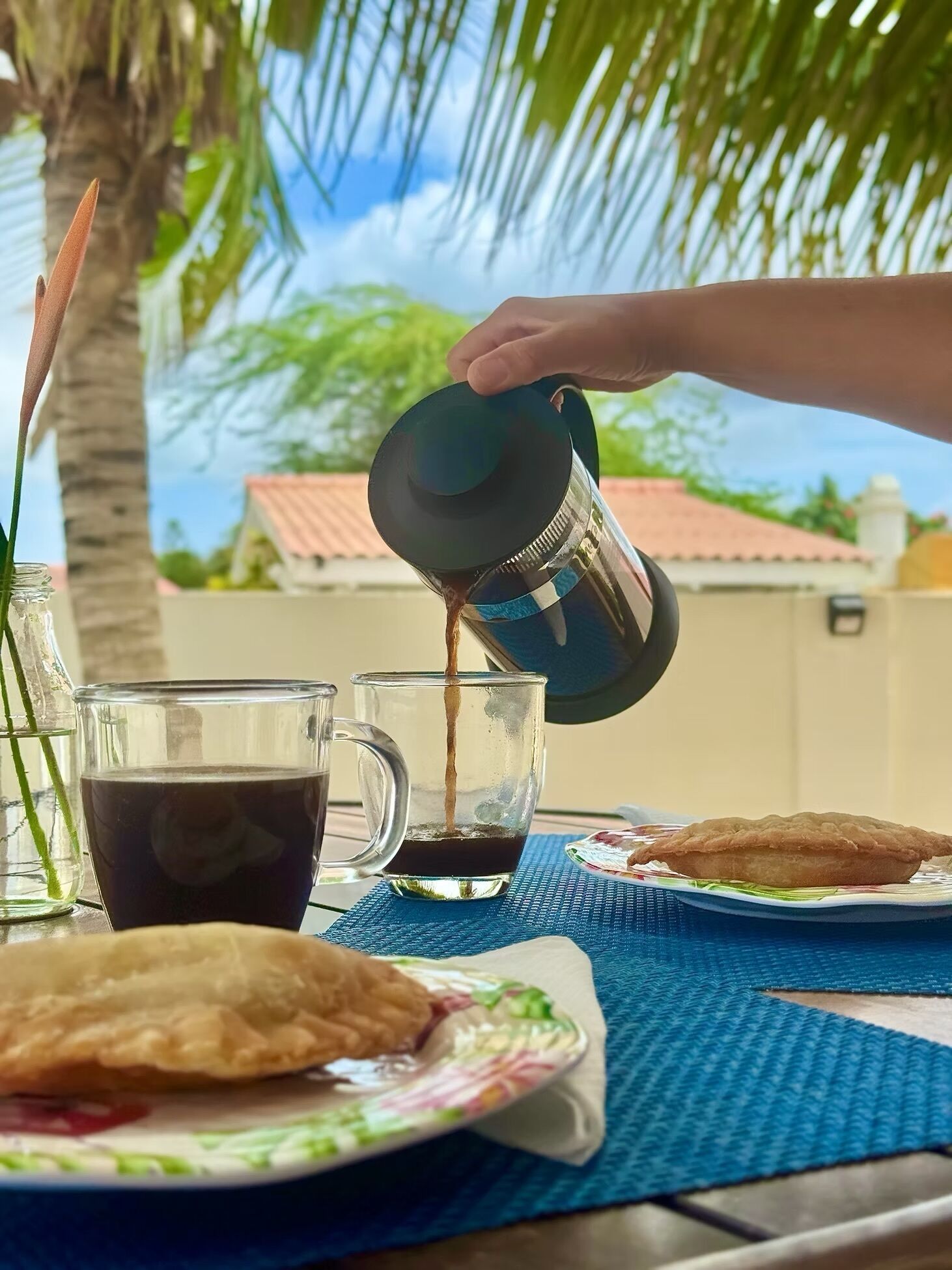 A hand pouring coffee into a glass mug, with a palm tree and a house in the background.