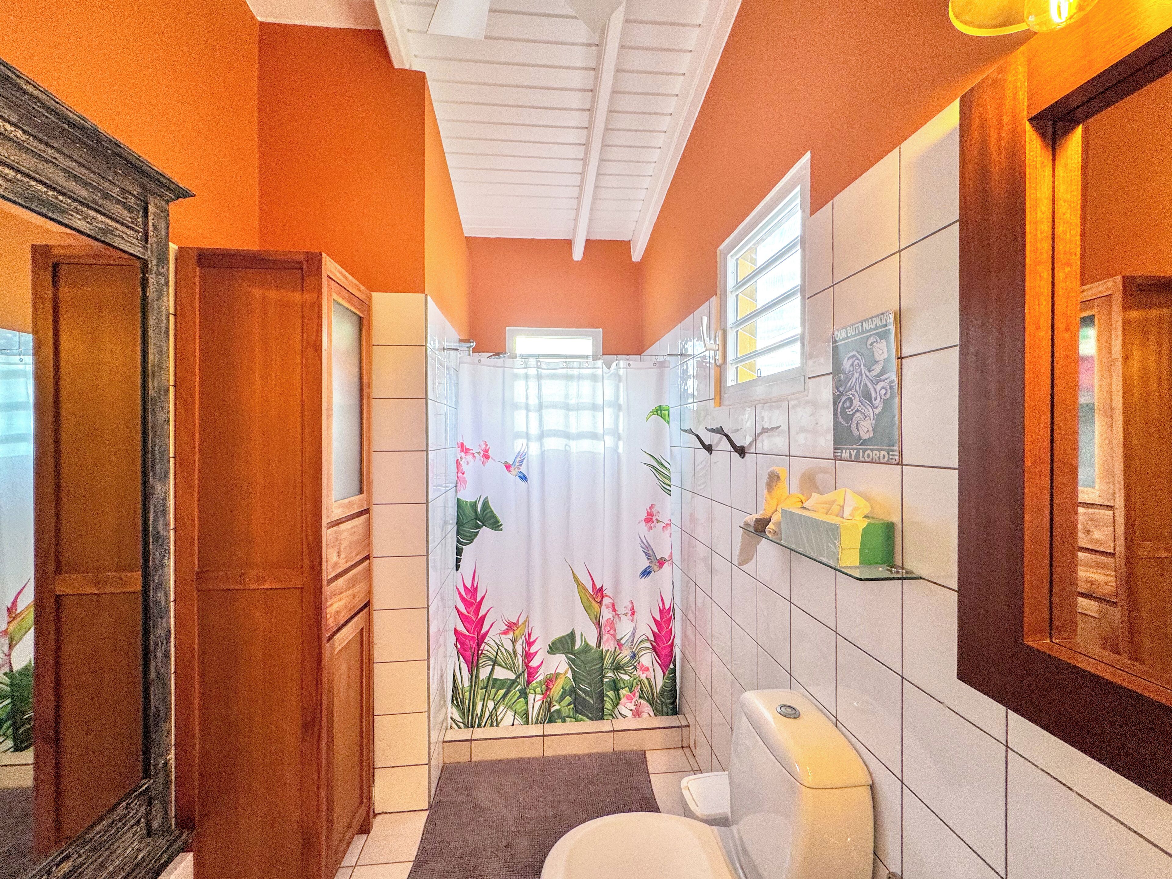 Un baño con cortina de ducha decorada con motivos florales tropicales, un inodoro y un espejo.