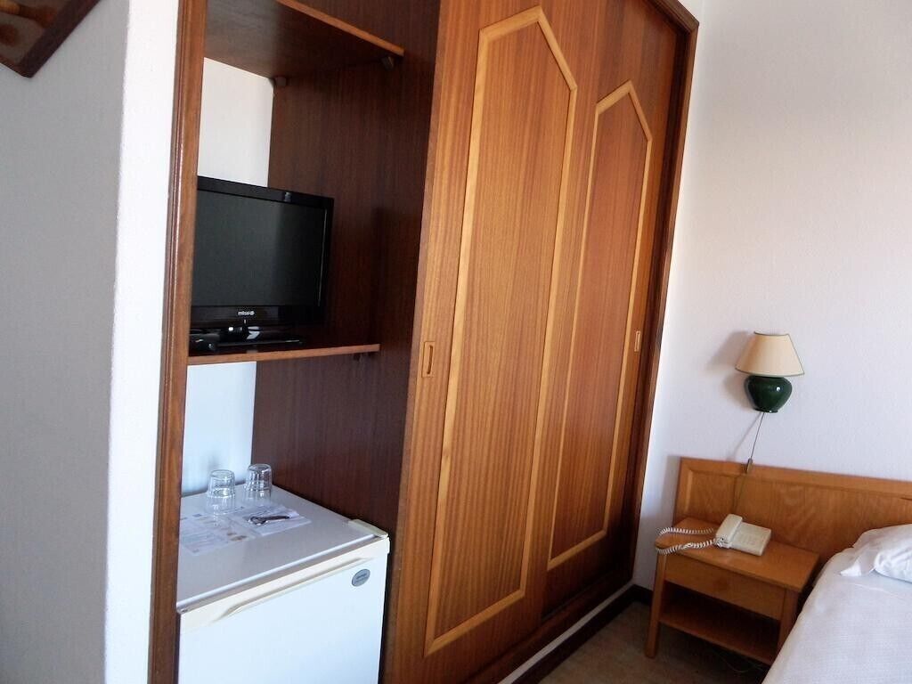 Habitación de hotel con un armario de madera, un televisor en una estantería, una mesita de noche con lámpara y una cama.