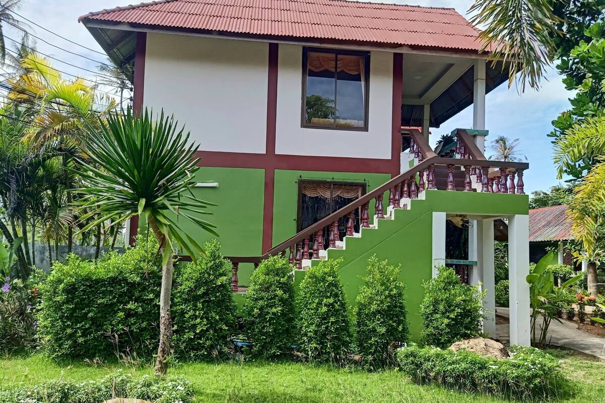Un edificio de dos pisos con techo rojo y detalles verdes, rodeado de exuberante vegetación y un jardín bien cuidado.