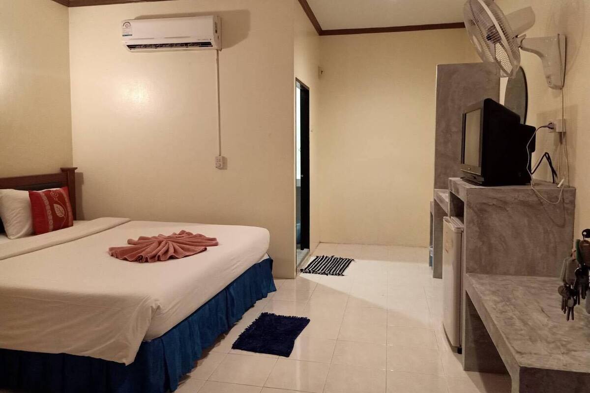Una habitación de hotel con cama, escritorio con computadora, aire acondicionado, ventilador de pared y una alfombra pequeña.