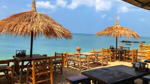 A beachside restaurant with wooden tables and chairs, thatched umbrellas, and a view of the ocean. - Blue Andaman Lanta Resort (Ko Lanta)