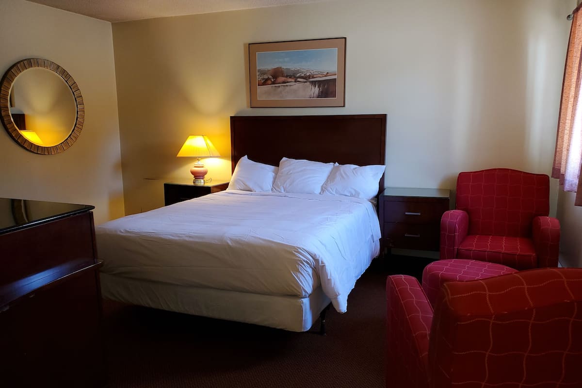 A hotel room with a large bed, a red armchair, a nightstand with a lamp, a mirror, and a framed picture on the wall.