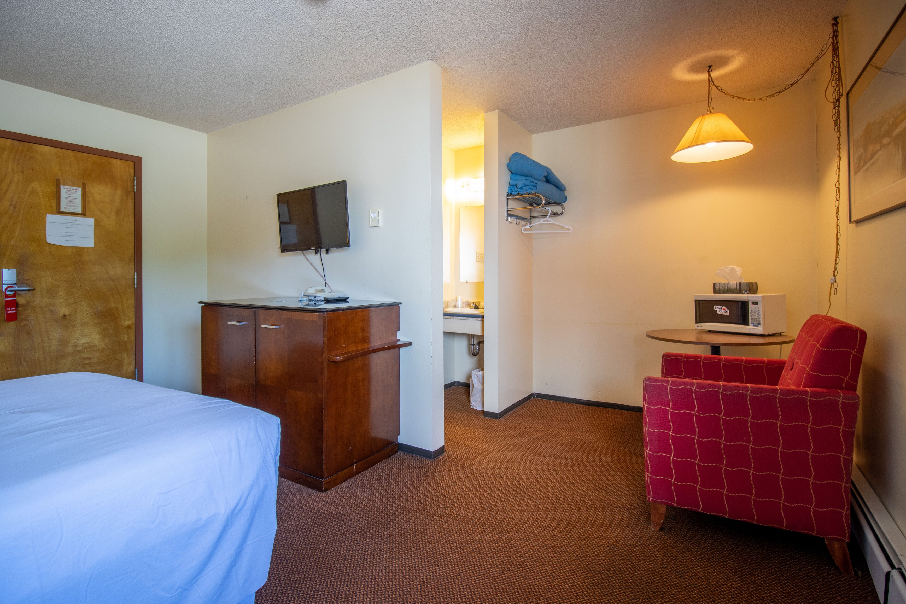 A hotel room with a bed, a desk with a computer, a chair, a TV, and a bathroom visible through an open door.