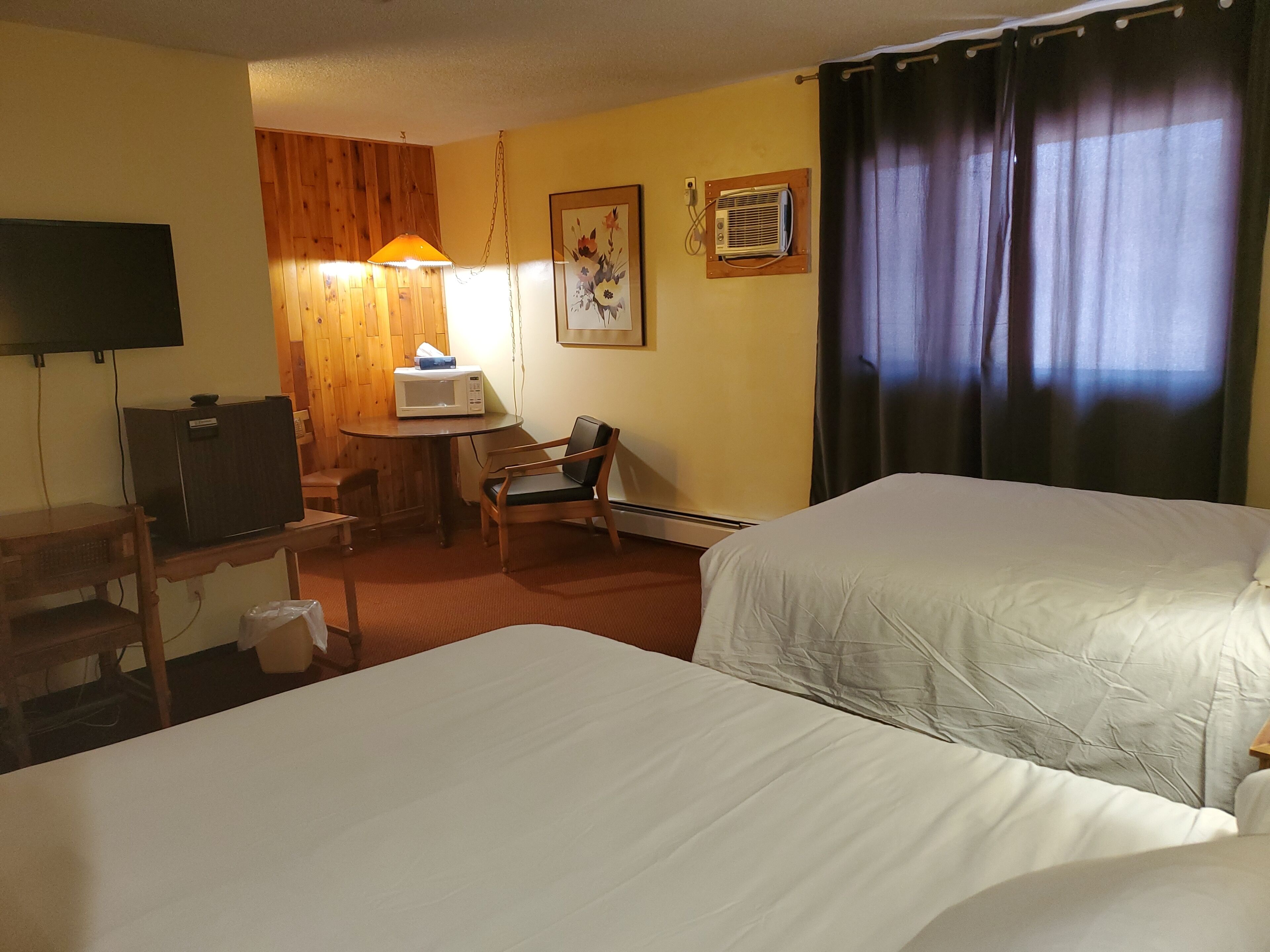 A hotel room with two beds, a desk with a computer, a chair, a small table with a microwave, a wall-mounted TV, and a window with curtains.