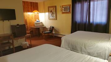 A hotel room with two beds, a desk with a computer, a chair, a small table with a microwave, a wall-mounted TV, and a window with curtains.