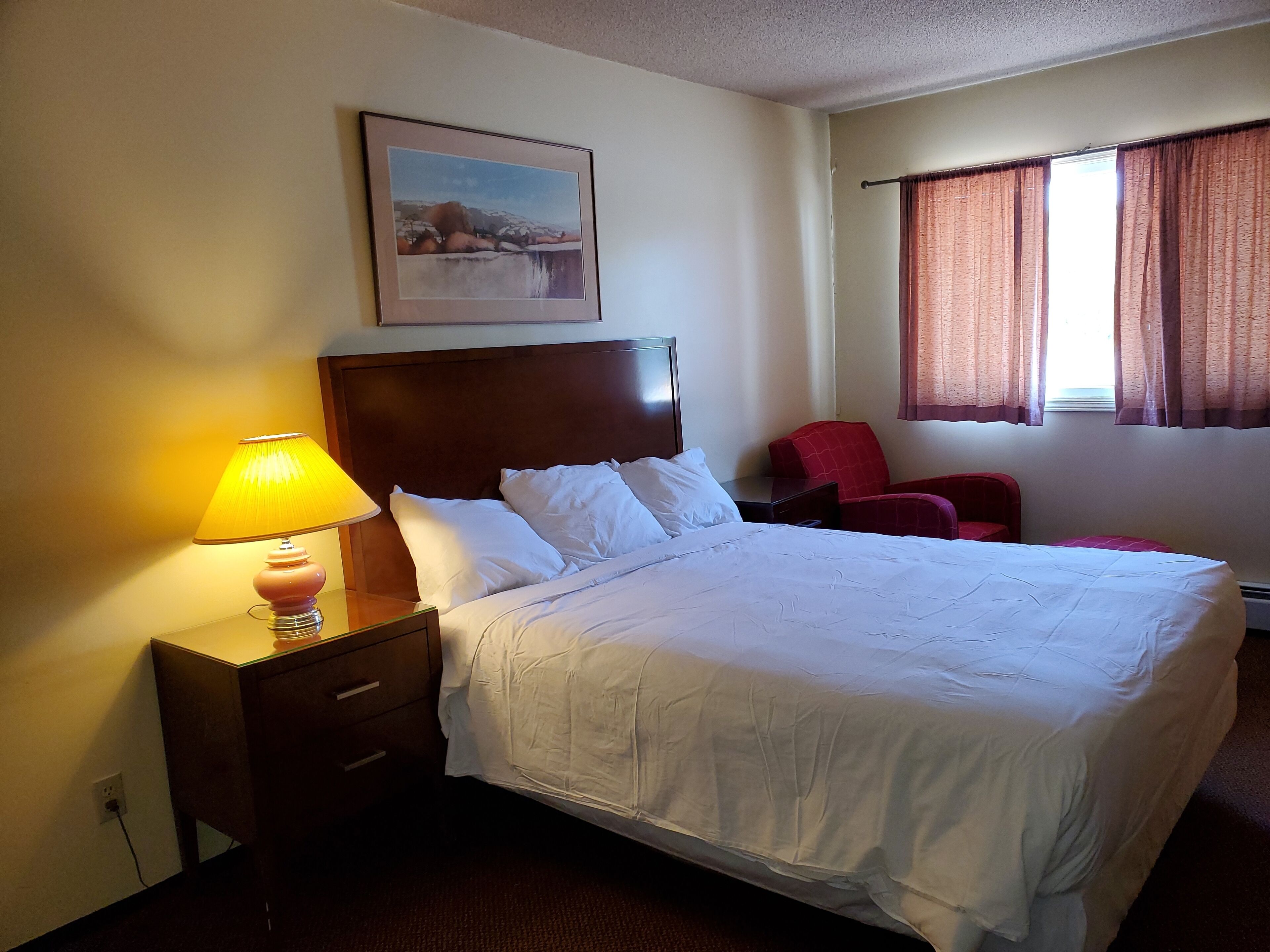 Habitación de hotel con cama, mesita de noche con lámpara, una silla roja, ventana con cortinas e imagen enmarcada en la pared.