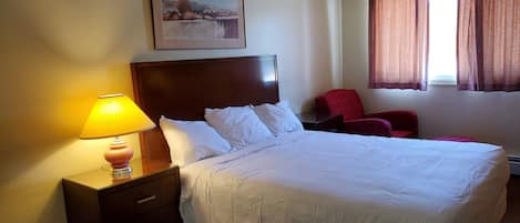 Habitación de hotel con cama, mesita de noche con lámpara, una silla roja, ventana con cortinas e imagen enmarcada en la pared.