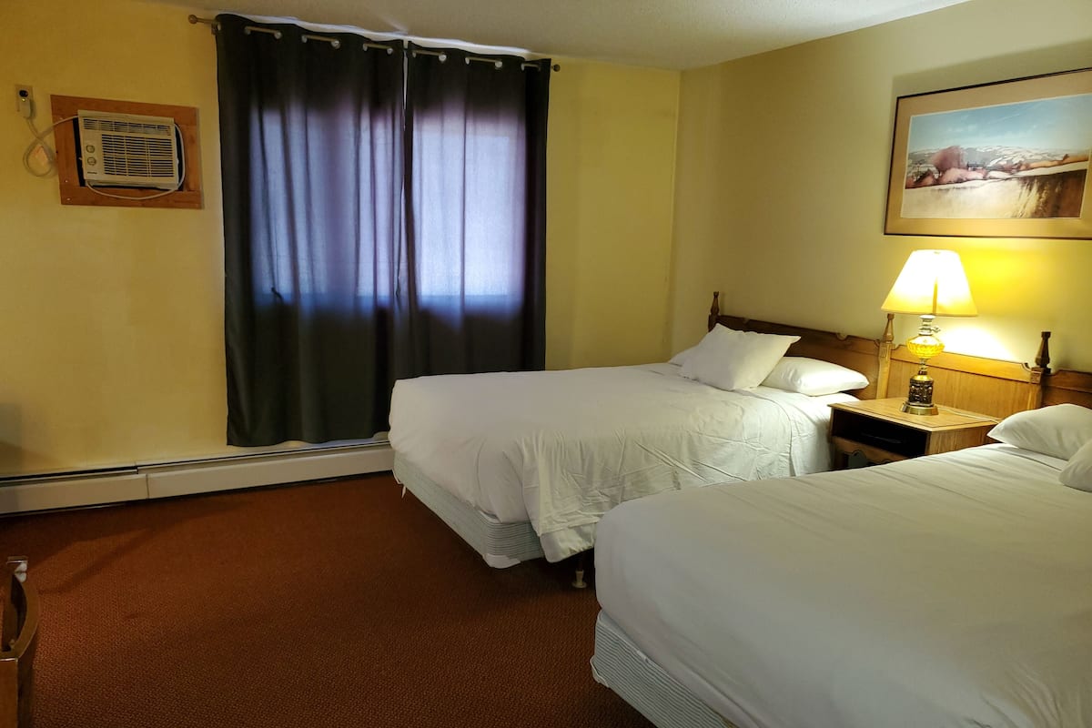 A hotel room with two beds, a nightstand with a lamp, a painting on the wall, and a window with curtains.