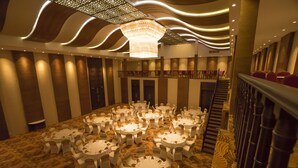A spacious banquet hall with multiple round tables and chairs, a central chandelier, and a staircase leading to an upper level. - Hotel Alakamanda (Anuradhapura)