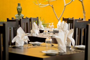 Mesa de jantar posta com guardanapos brancos, copos transparentes e uma estátua de Buda ao fundo.