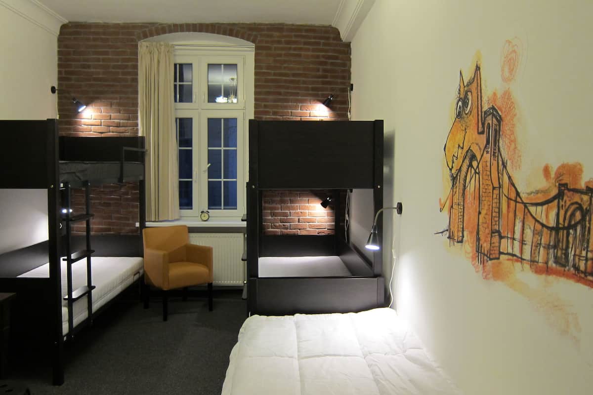 A dormitory room with bunk beds, a small chair, a radiator, a window with curtains, and a mural on the wall.
