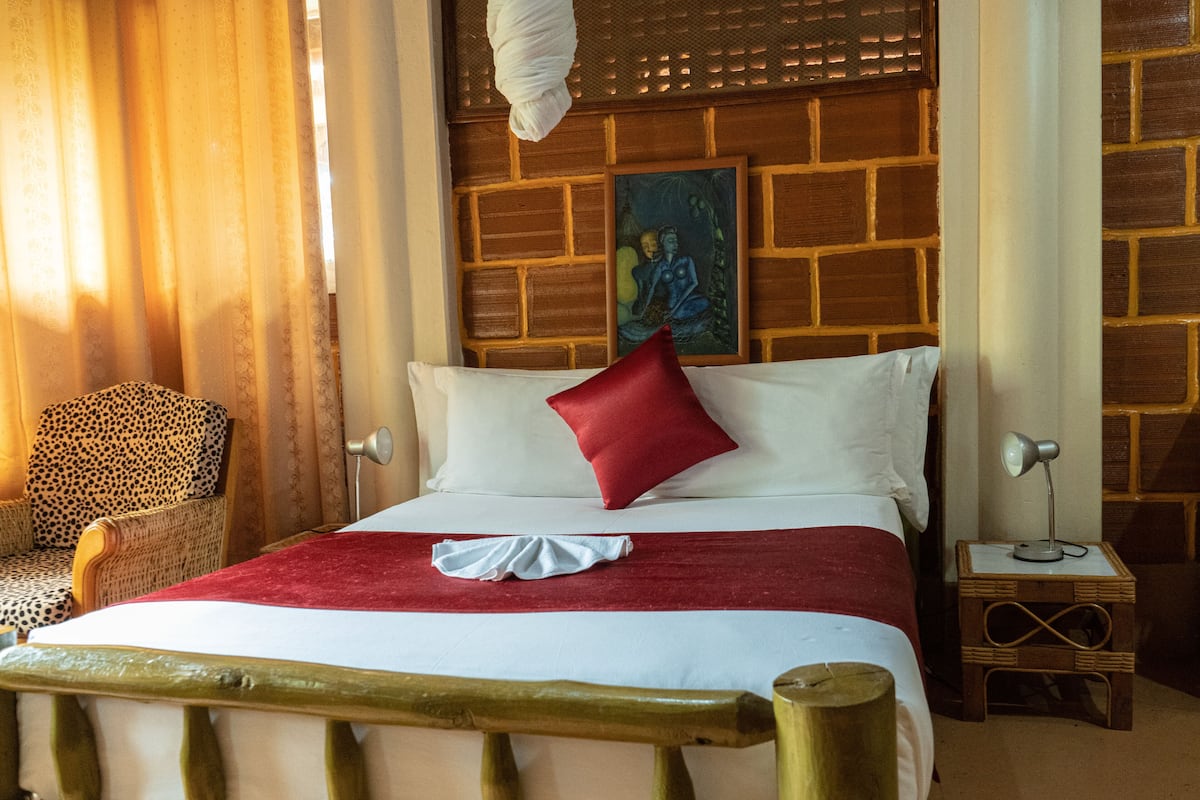 A bed with a red pillow, white linens, and a wooden headboard. There is a chair with a leopard print cushion, a small table with a lamp, and a painting on the wall.