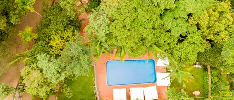 An aerial view of a property with a swimming pool surrounded by lush greenery and a paved area with outdoor furniture.