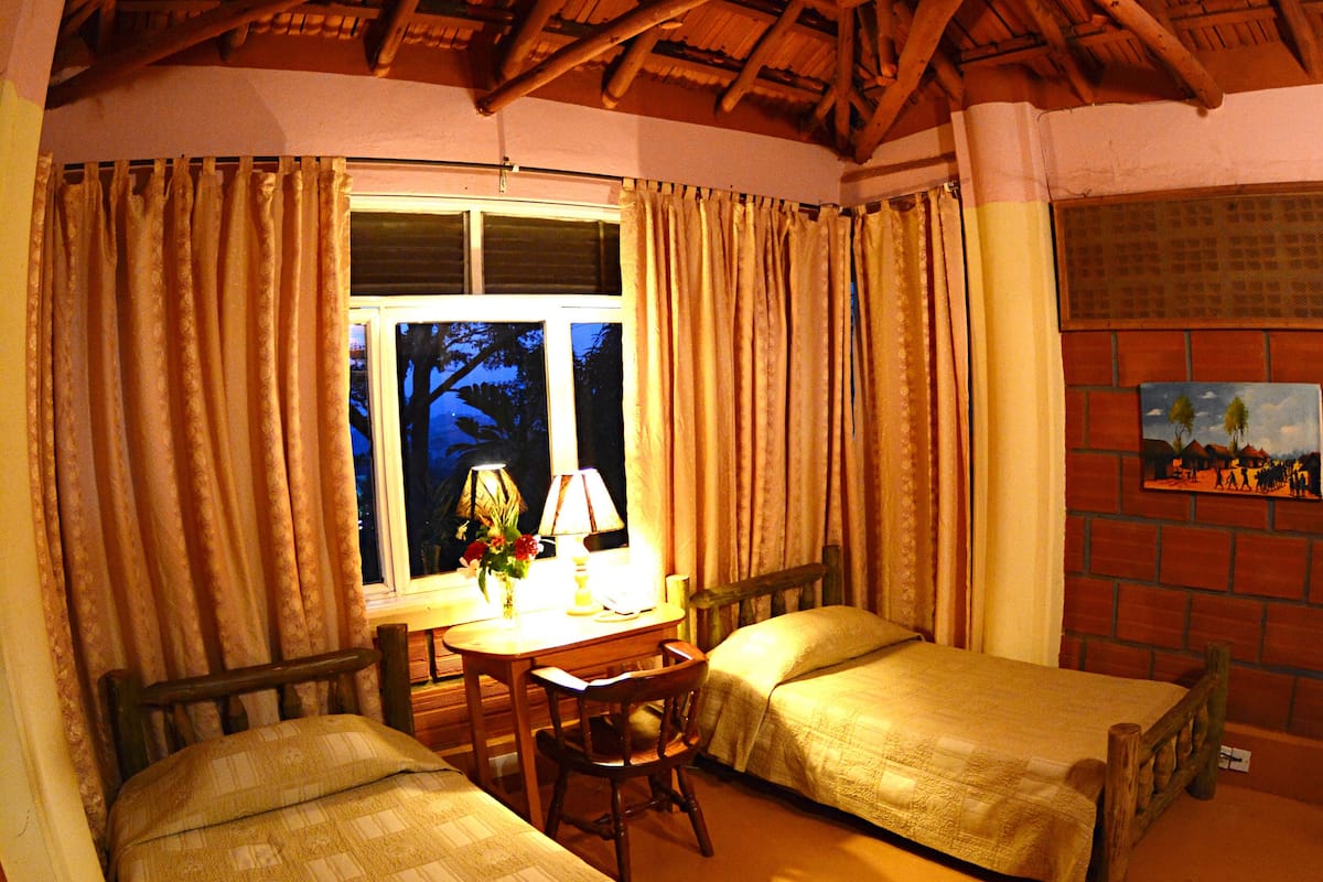 A room with two wooden beds, a small table with a lamp, and a window with curtains.