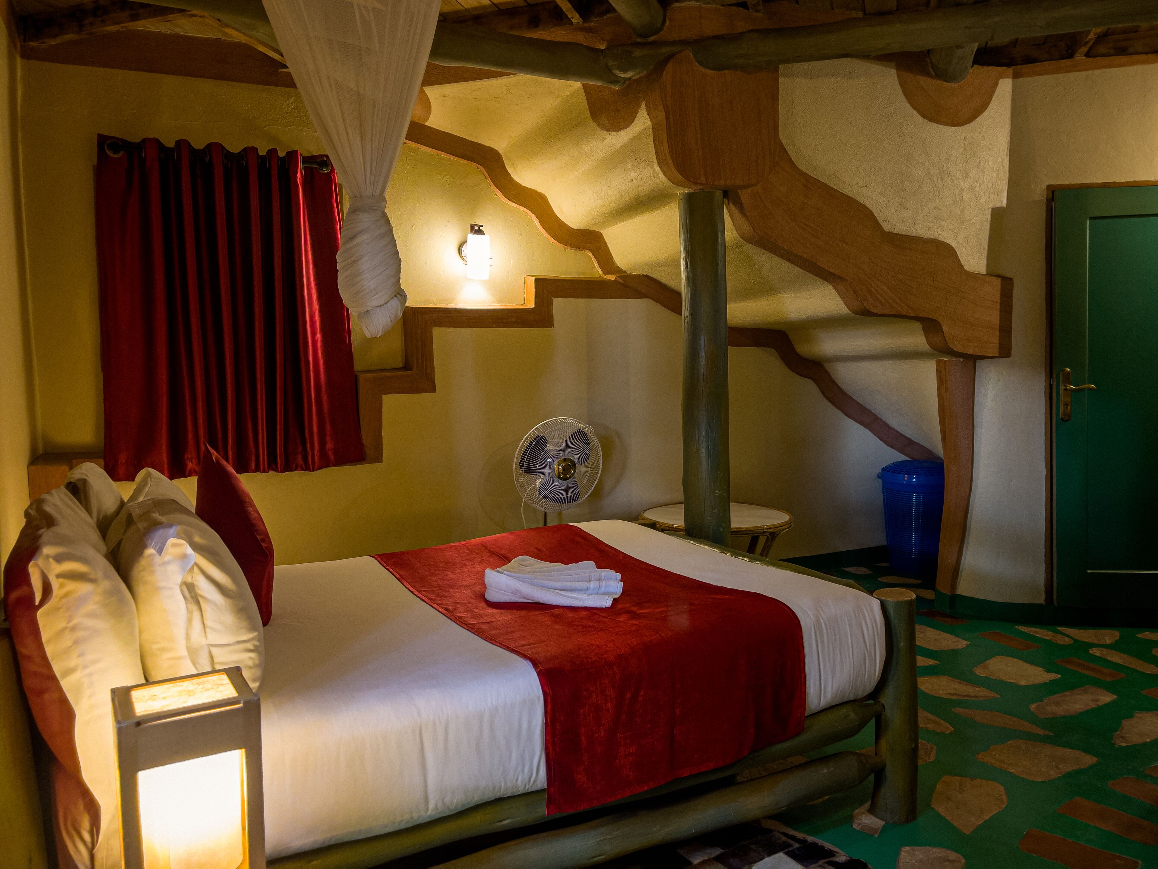 A bed with a red blanket, white pillows, and a bedside lamp. There is a wall-mounted fan and a green door in the room.