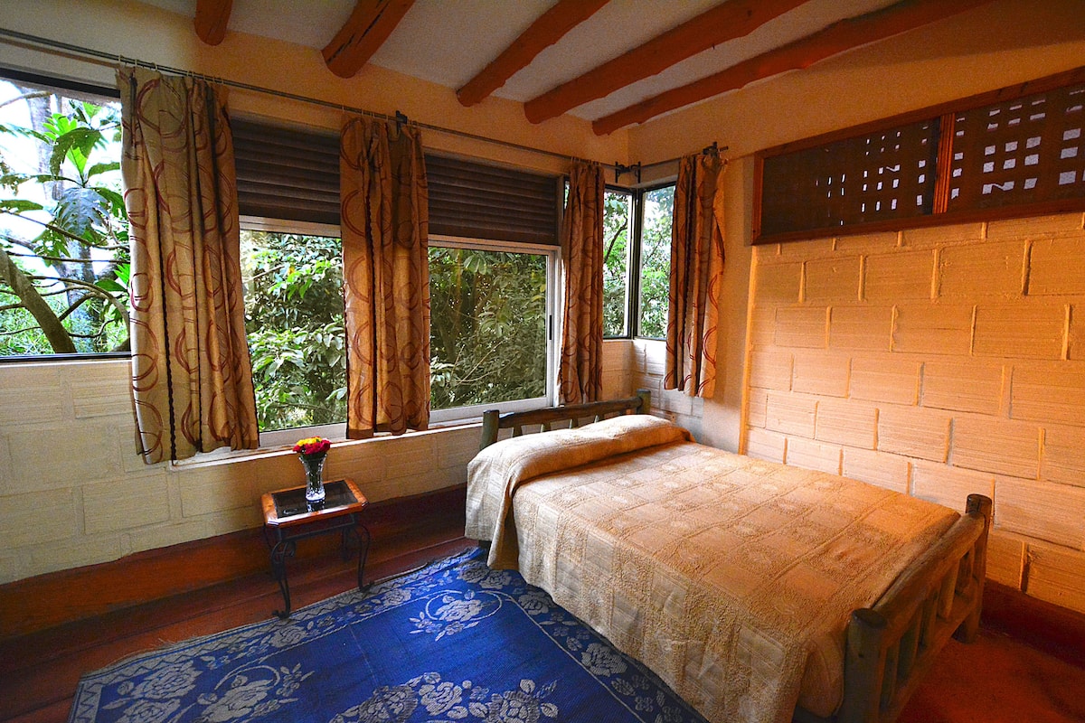A bedroom with a bed, a window with curtains, a small table with a vase, and a blue patterned rug.
