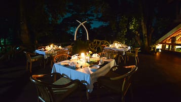 Un área de comedor al aire libre con mesas dispuestas para una comida, rodeada de sillas de madera y un arco decorativo.