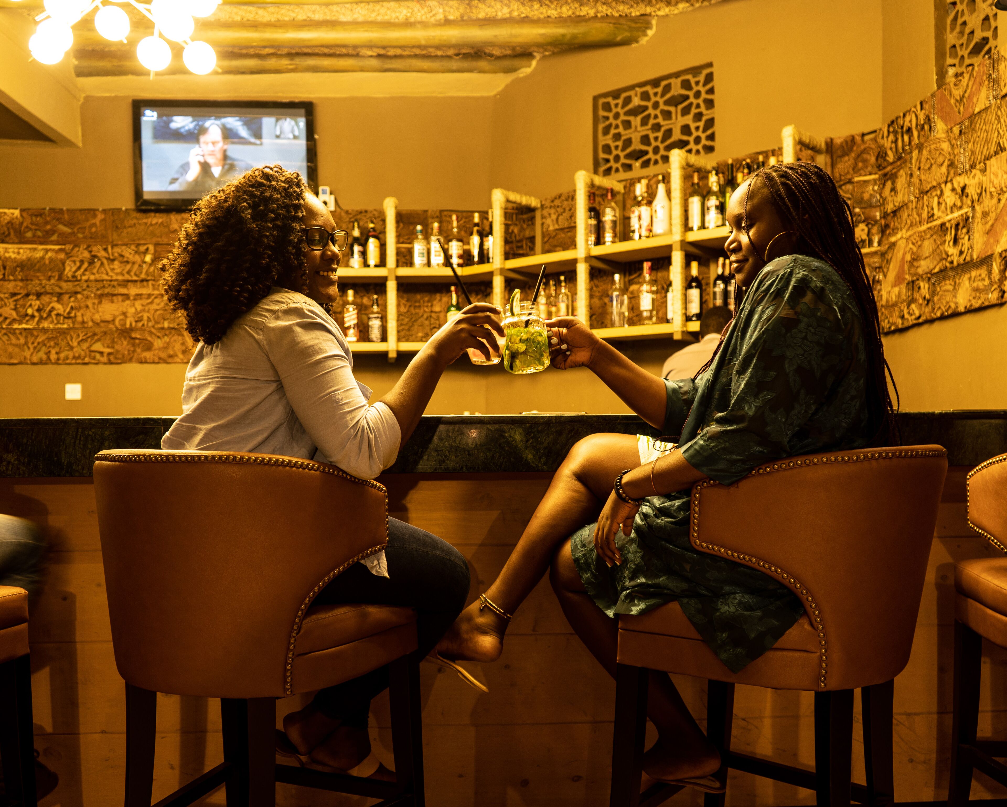 Two people seated at a bar with a television in the background and shelves of bottles.