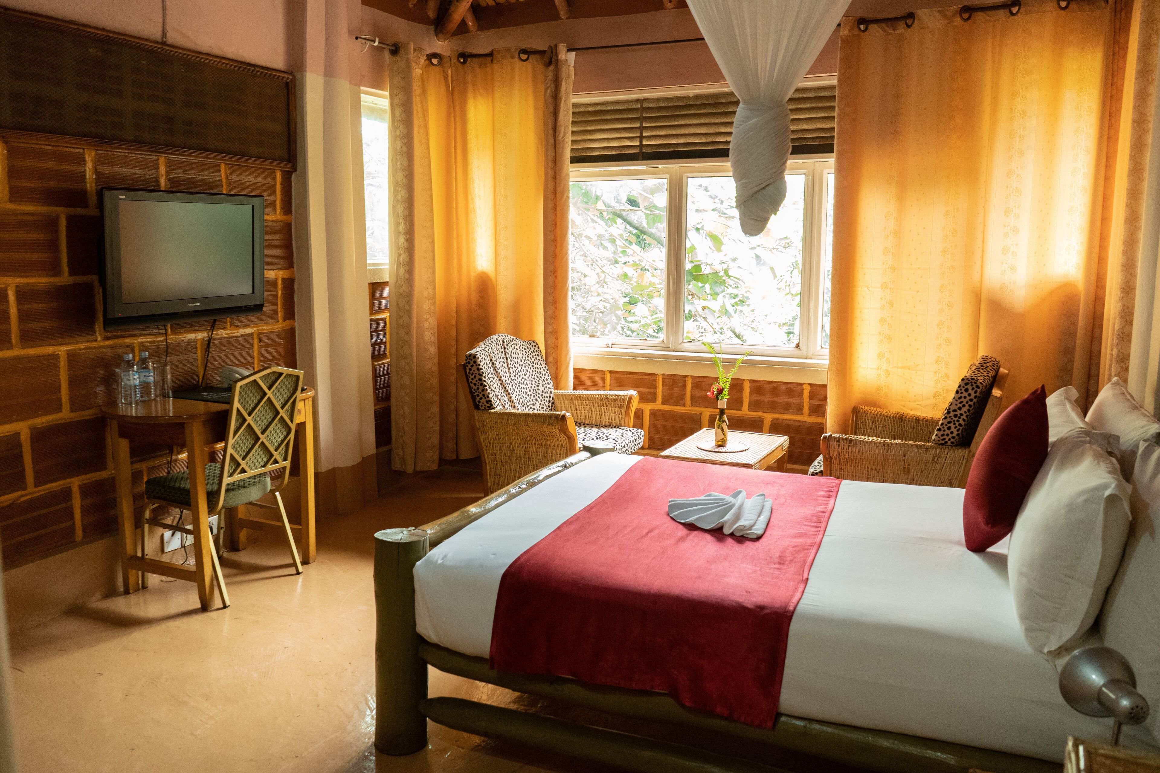 A hotel room with a bed, a TV, wicker furniture, a window with curtains, and a view of greenery outside.