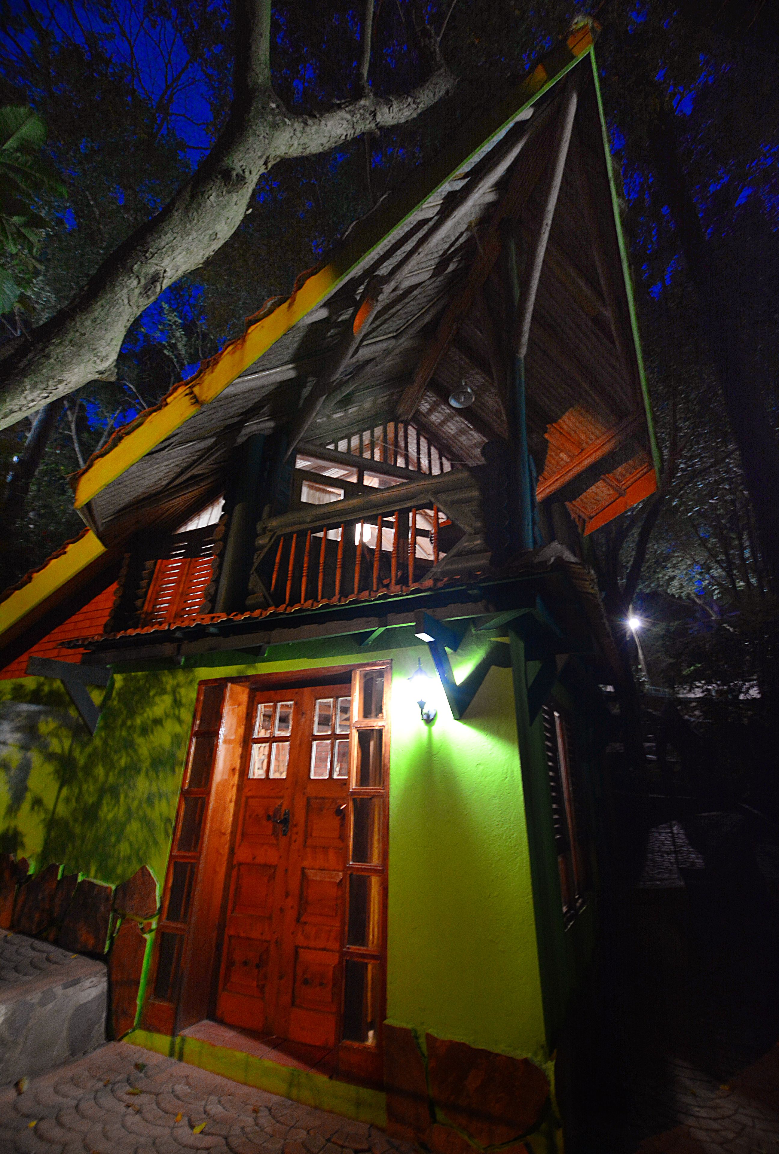 Une maison en bois à deux étages, à l’extérieur vert et à la porte rouge, entourée d’arbres.