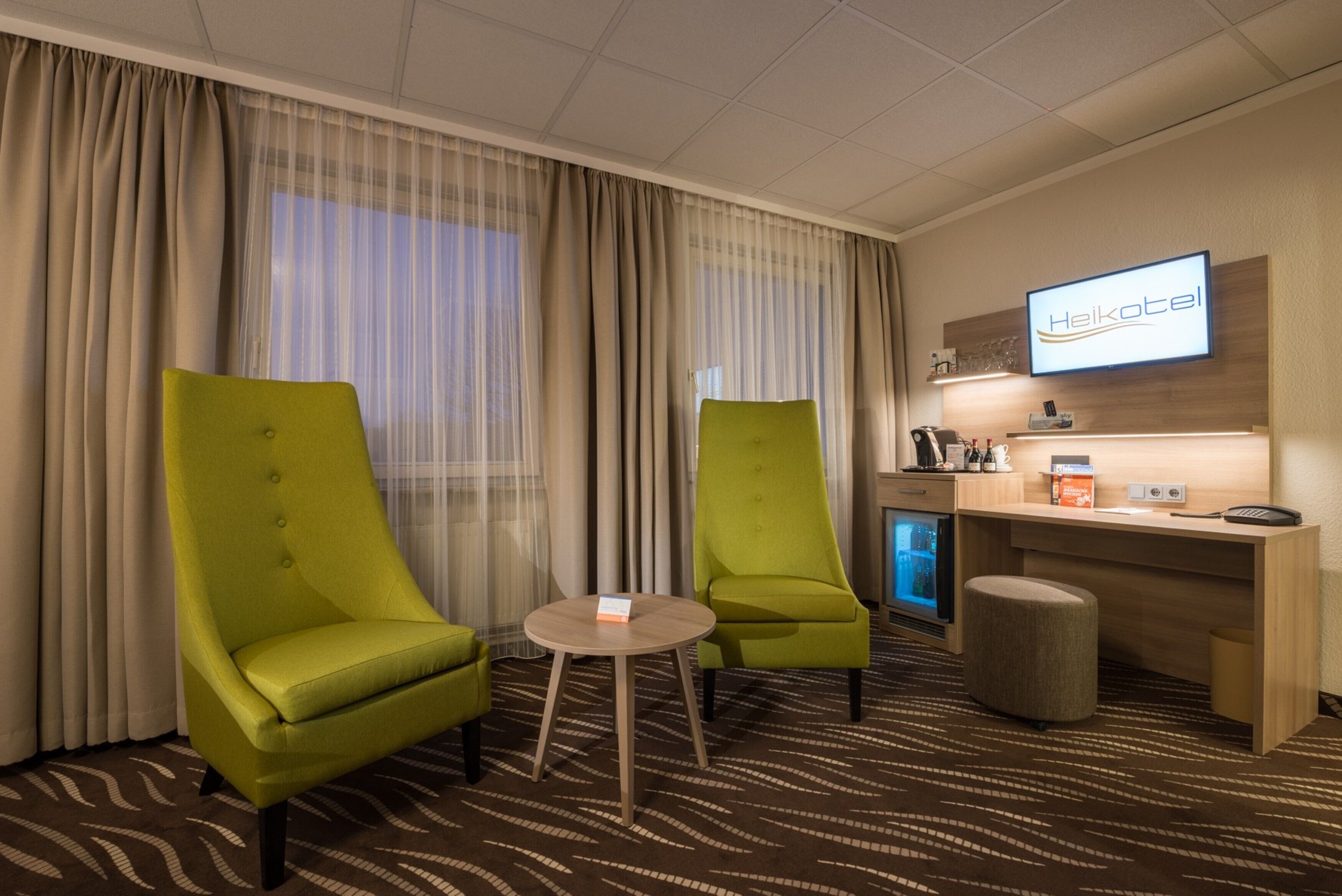 Ein Hotelzimmer mit zwei grünen Sesseln, einem kleinen runden Tisch, einem Schreibtisch mit Computer und einem Fernseher, der den Namen des Hotels anzeigt.