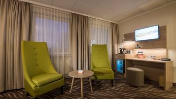 Ein Hotelzimmer mit zwei grünen Sesseln, einem kleinen runden Tisch, einem Schreibtisch mit Computer und einem Fernseher, der den Namen des Hotels anzeigt.
