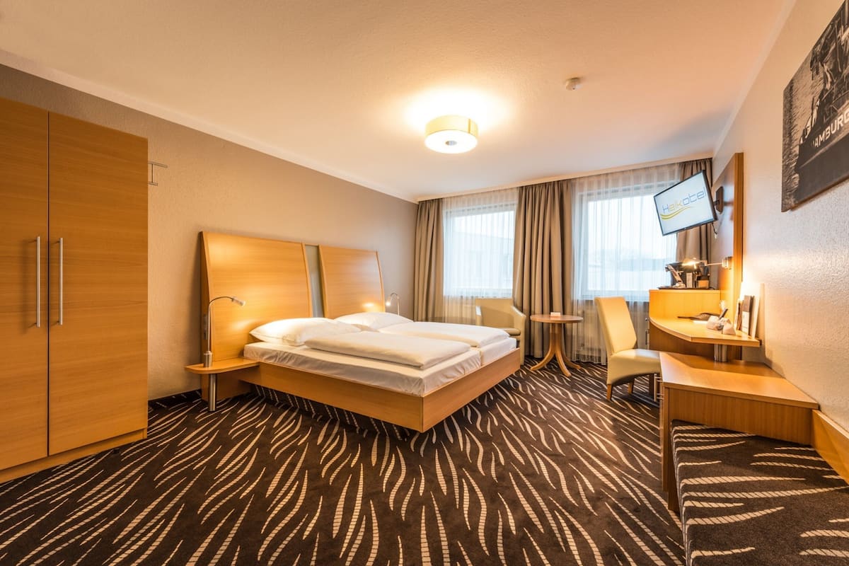 Quarto de hotel com cama, escrivaninha, cadeira, televisão e guarda-roupa.
