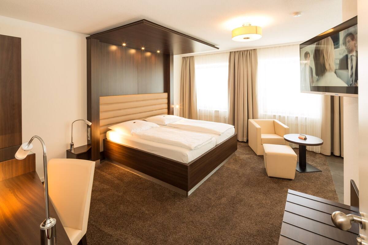 Quarto de hotel moderno com uma cama grande, área de estar com uma poltrona e uma mesinha, TV fixada na parede e uma janela ampla com cortinas.