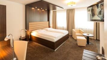 Ein modernes Hotelzimmer mit einem großen Bett, einer Sitzecke mit einem Sessel und einem kleinen Tisch, einem an der Wand montierten Fernseher und einem großen Fenster mit Vorhängen.