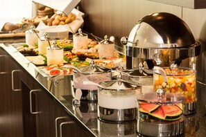 Un bufé con diversos alimentos y bebidas, que incluye frutas, ensaladas y platos calientes, servidos en recipientes metálicos con tapas.