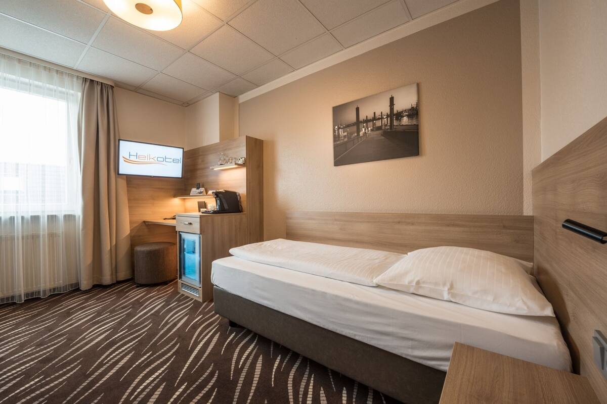 Quarto de hotel com cama, escrivaninha, televisão e pintura de paisagem urbana na parede.