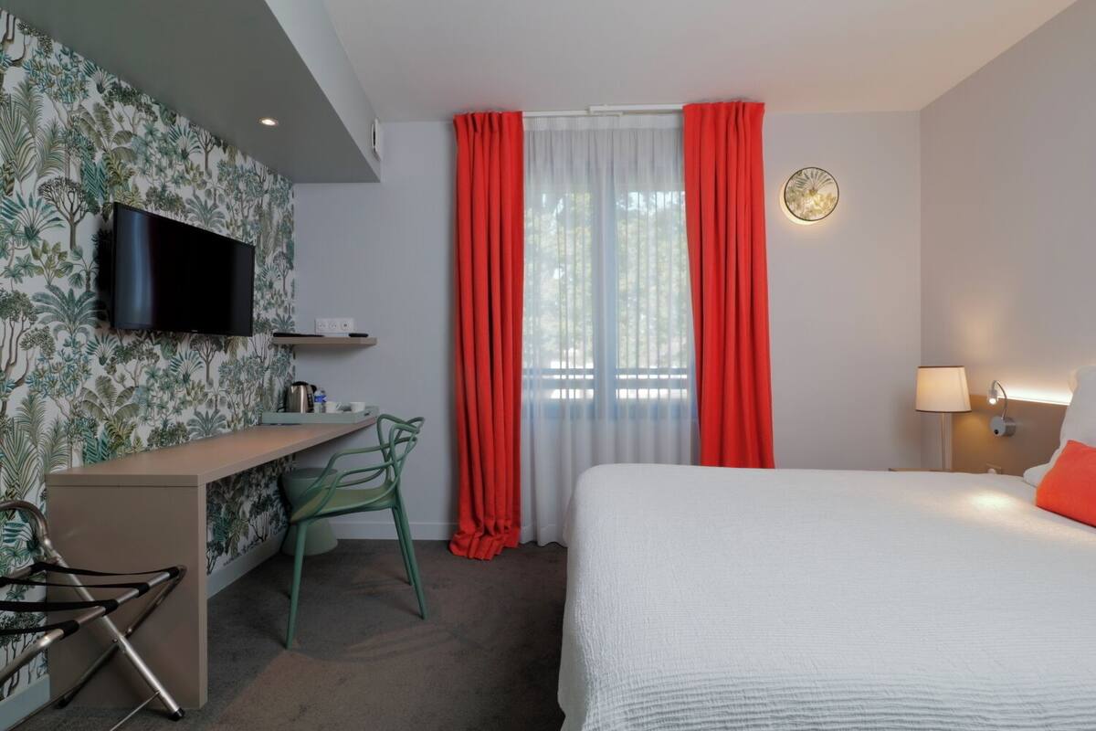 Habitación de hotel con cama, escritorio con silla, televisor y ventana con cortinas rojas.