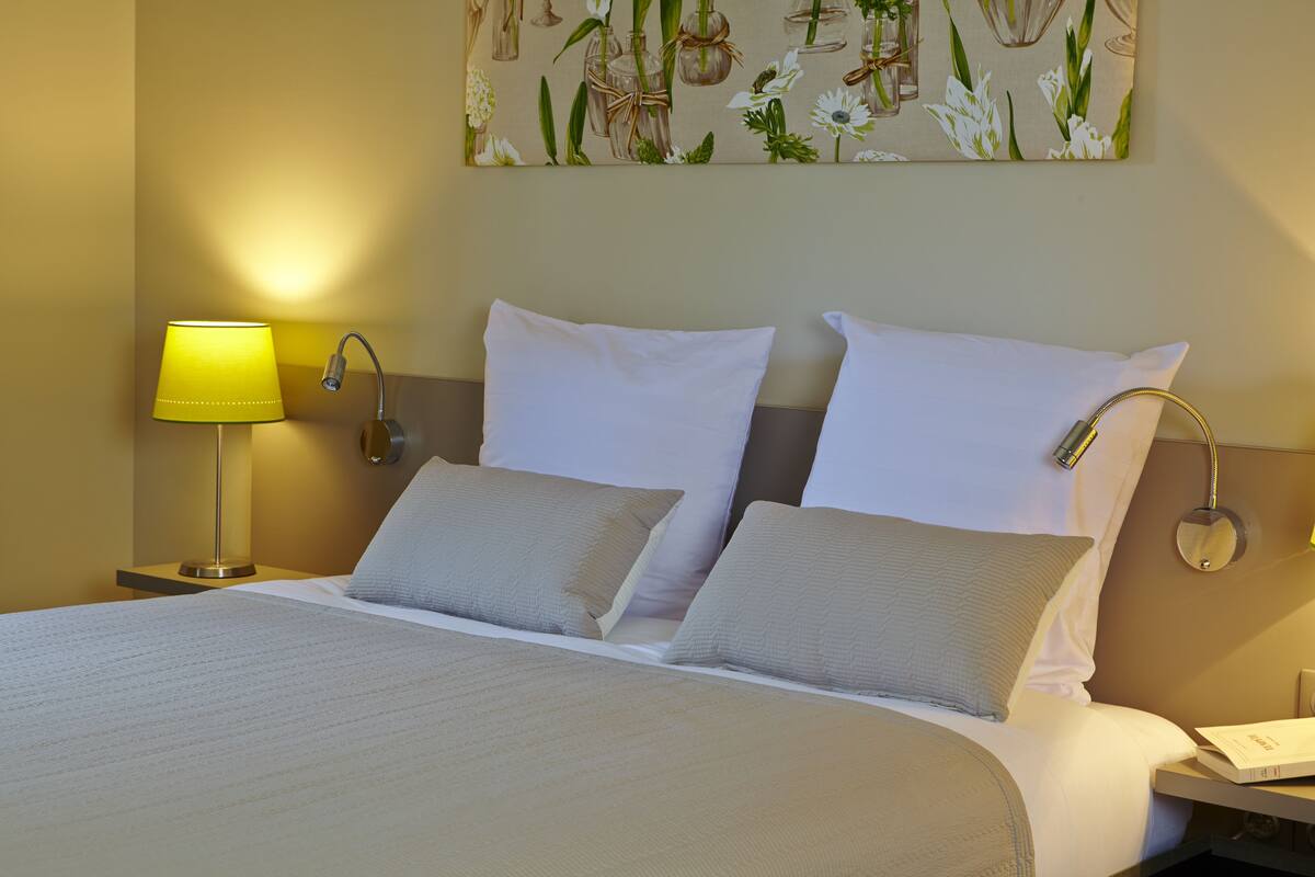 Una cama bien hecha con sábanas blancas y dos almohadas, una lámpara de noche y un espejo montado en la pared.