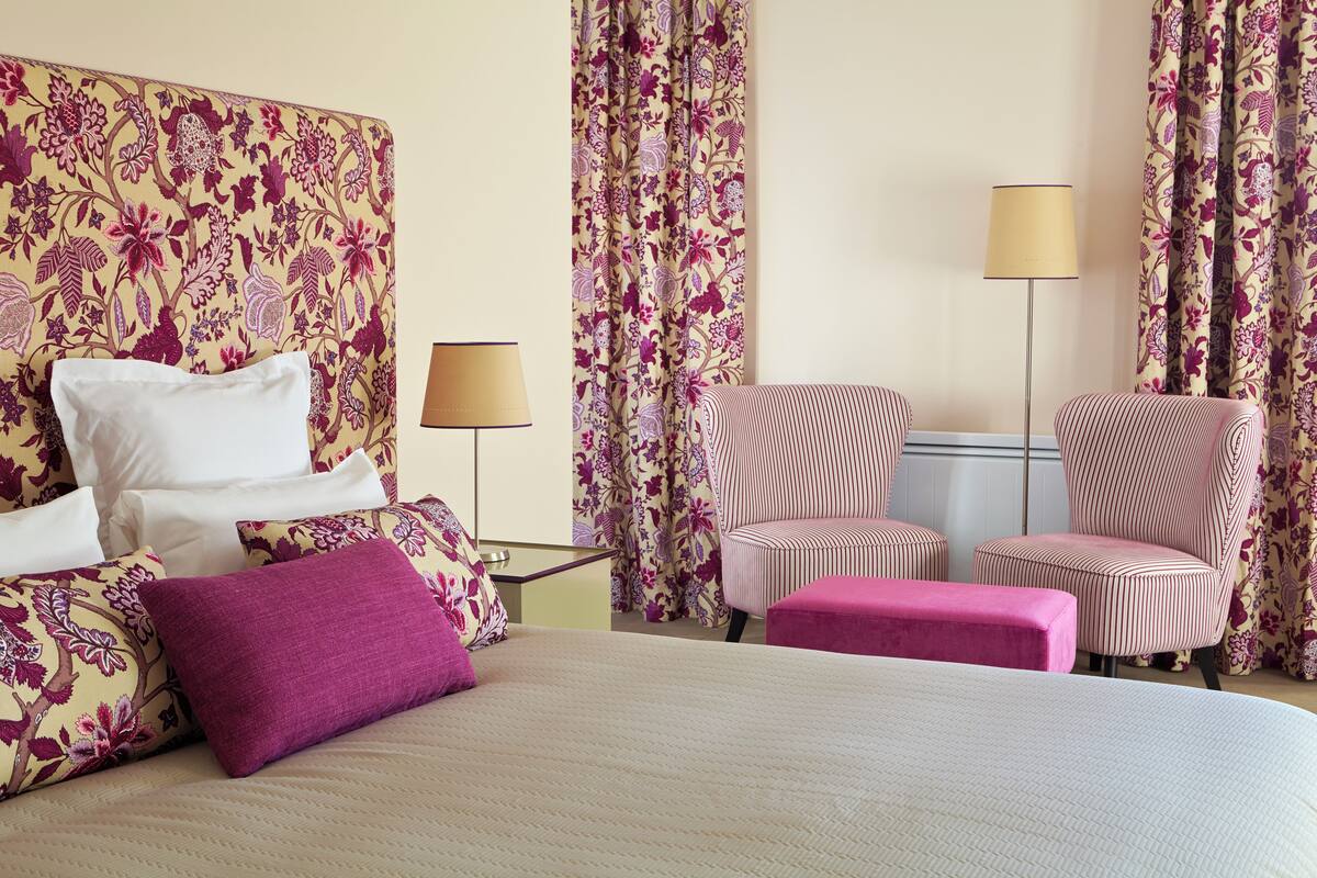 Habitación de hotel con cama, dos sillas, un banco y cortinas con estampado floral.