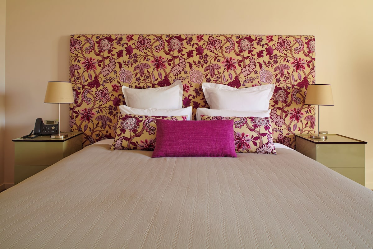 Una cama bien tendida con ropa de cama blanca y morada, dos lámparas de noche y un cabecero estampado con motivos florales.
