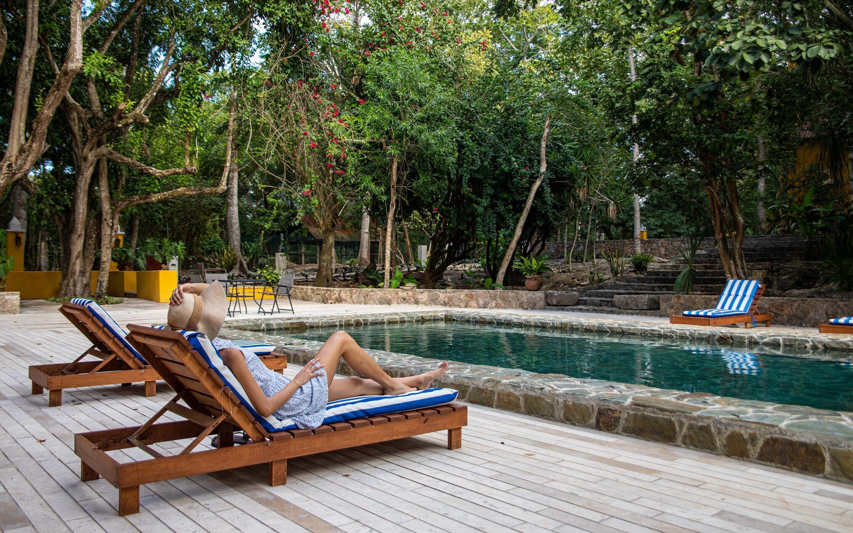 Un área junto a la piscina con sillones de madera, una piscina de piedra y abundante vegetación.