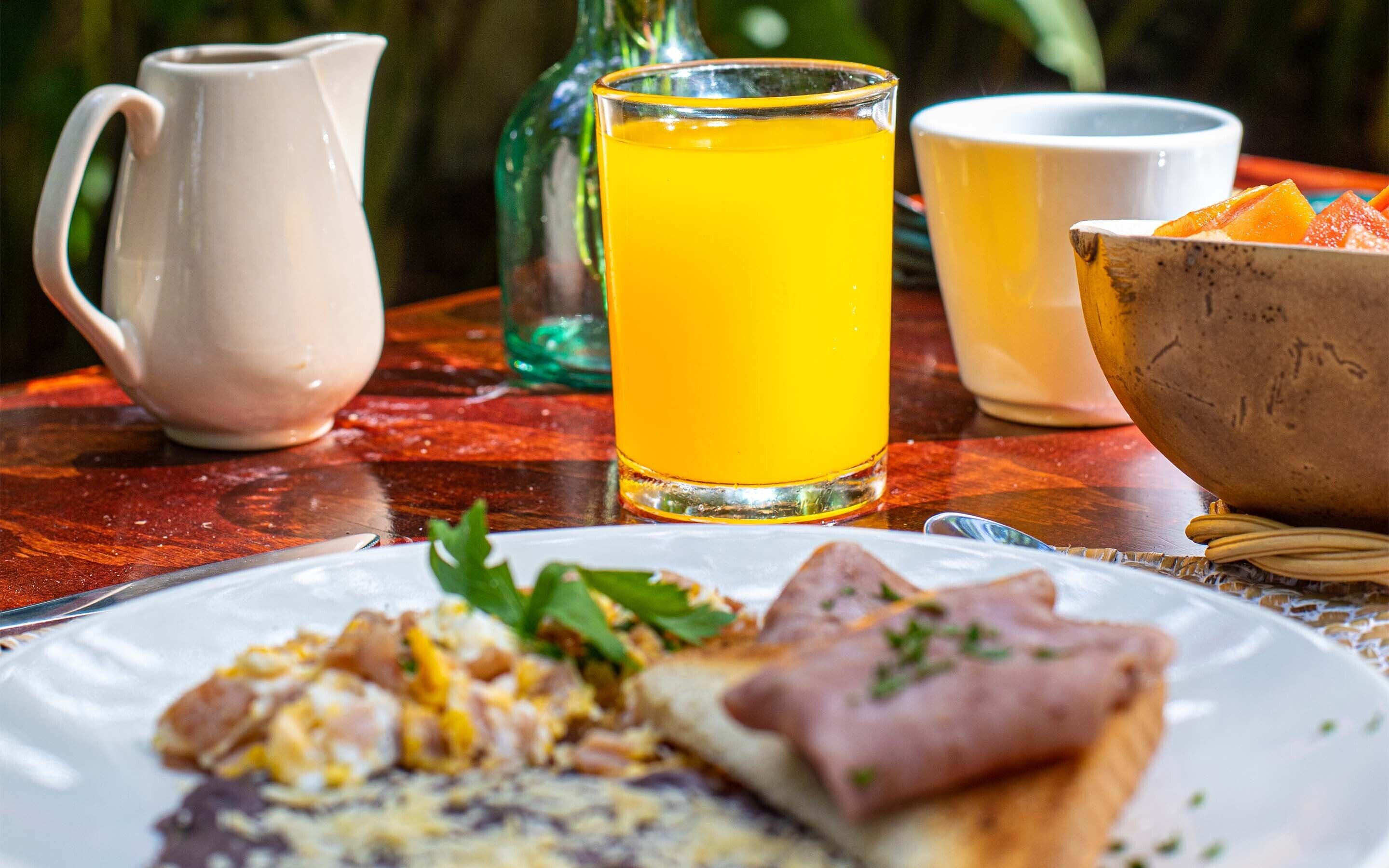 Un desayuno con un plato de comida, una jarra, un vaso de zumo de naranja y un bol de fruta.