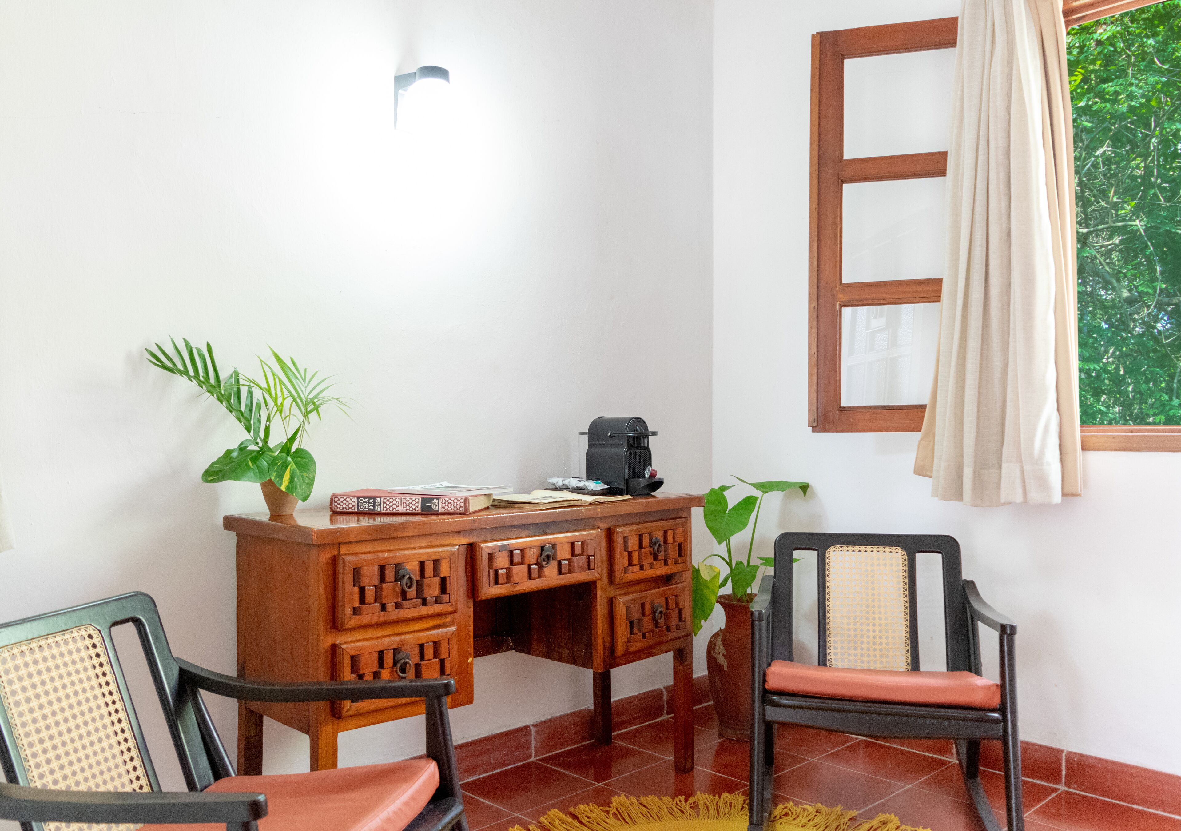 Una habitación con un escritorio de madera, dos sillas, una planta en maceta y una ventana con cortina.