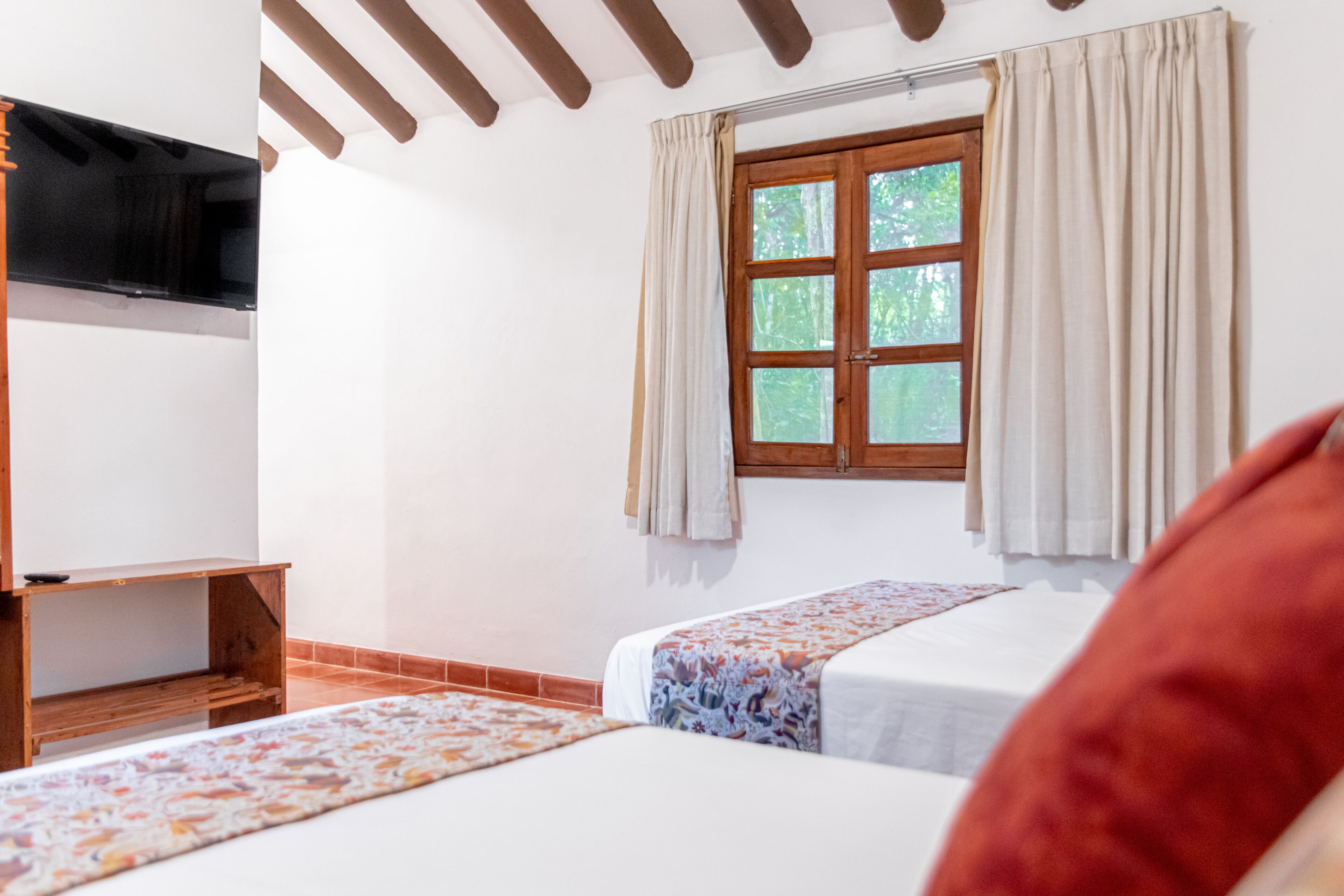 Une chambre avec deux lits, un meuble TV en bois, une fenêtre avec des rideaux et un sol carrelé.