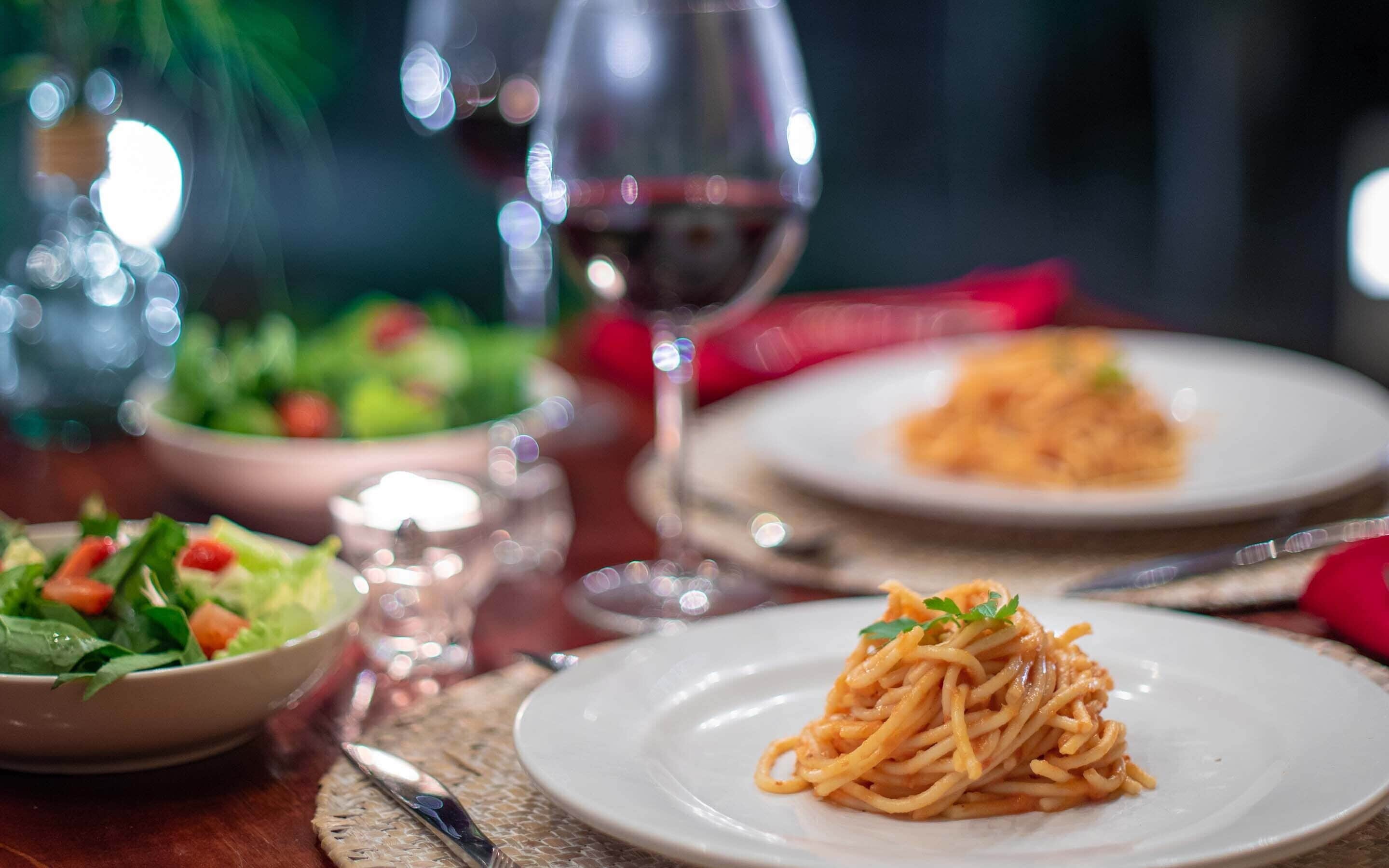 A dining table with plates of spaghetti and salad, a wine glass, and cutlery.