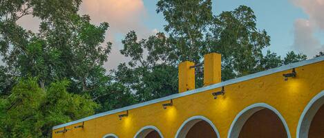 Un edificio amarillo con arcos blancos y un balcón, rodeado de vegetación.