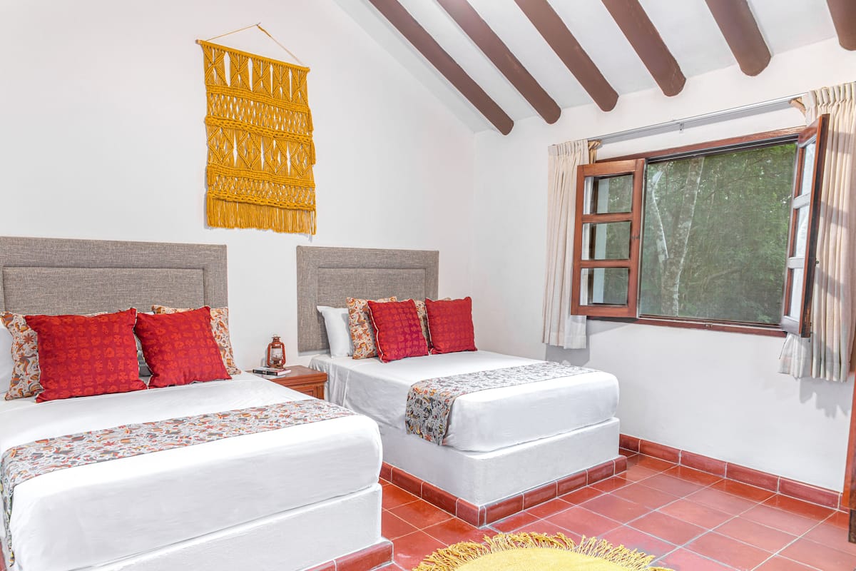 A hotel room with two beds, a large window with curtains, a macramé wall hanging, and a tiled floor.