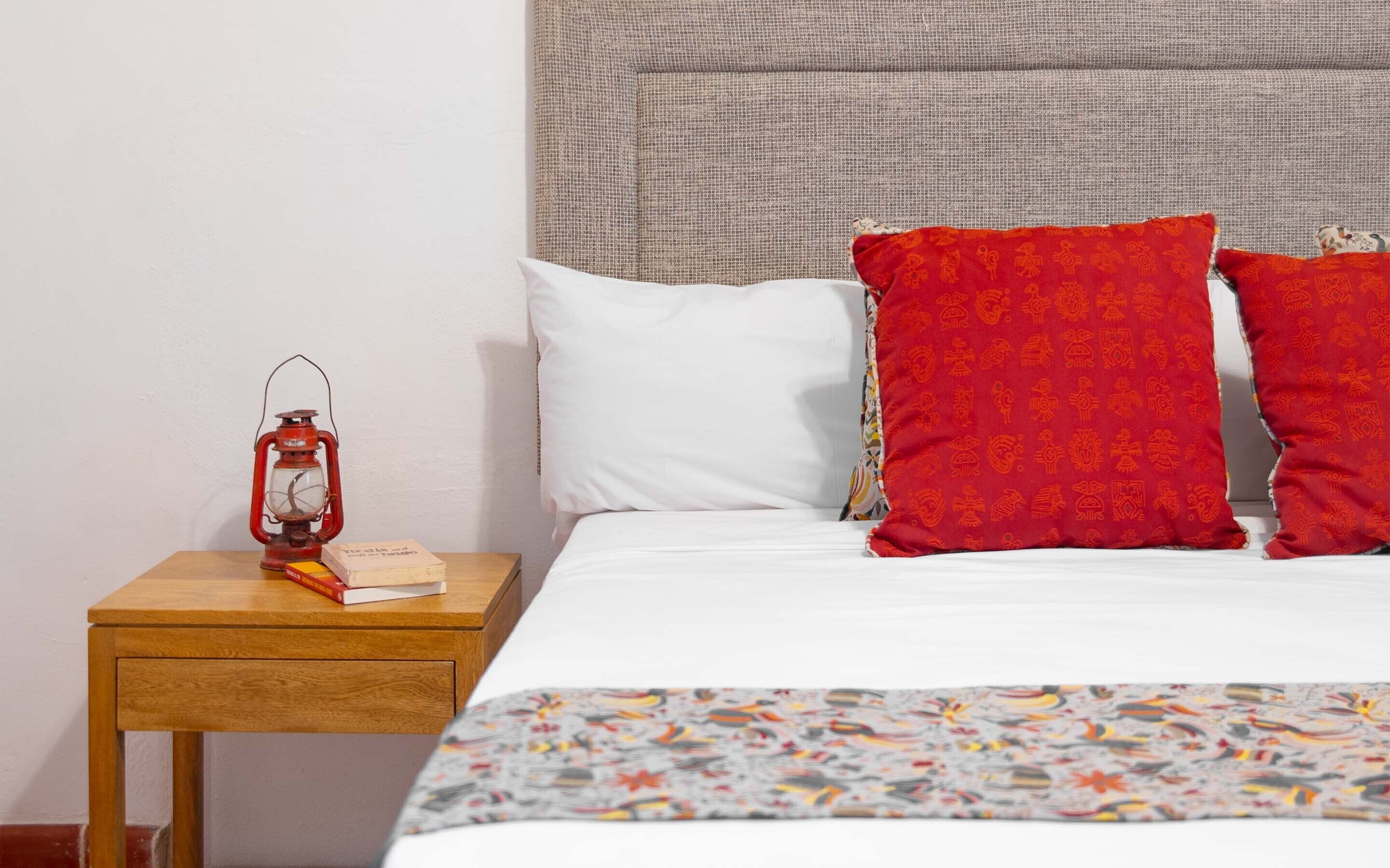 A neatly made bed with white and red pillows, a patterned blanket, and a small wooden nightstand with a red lantern and books.