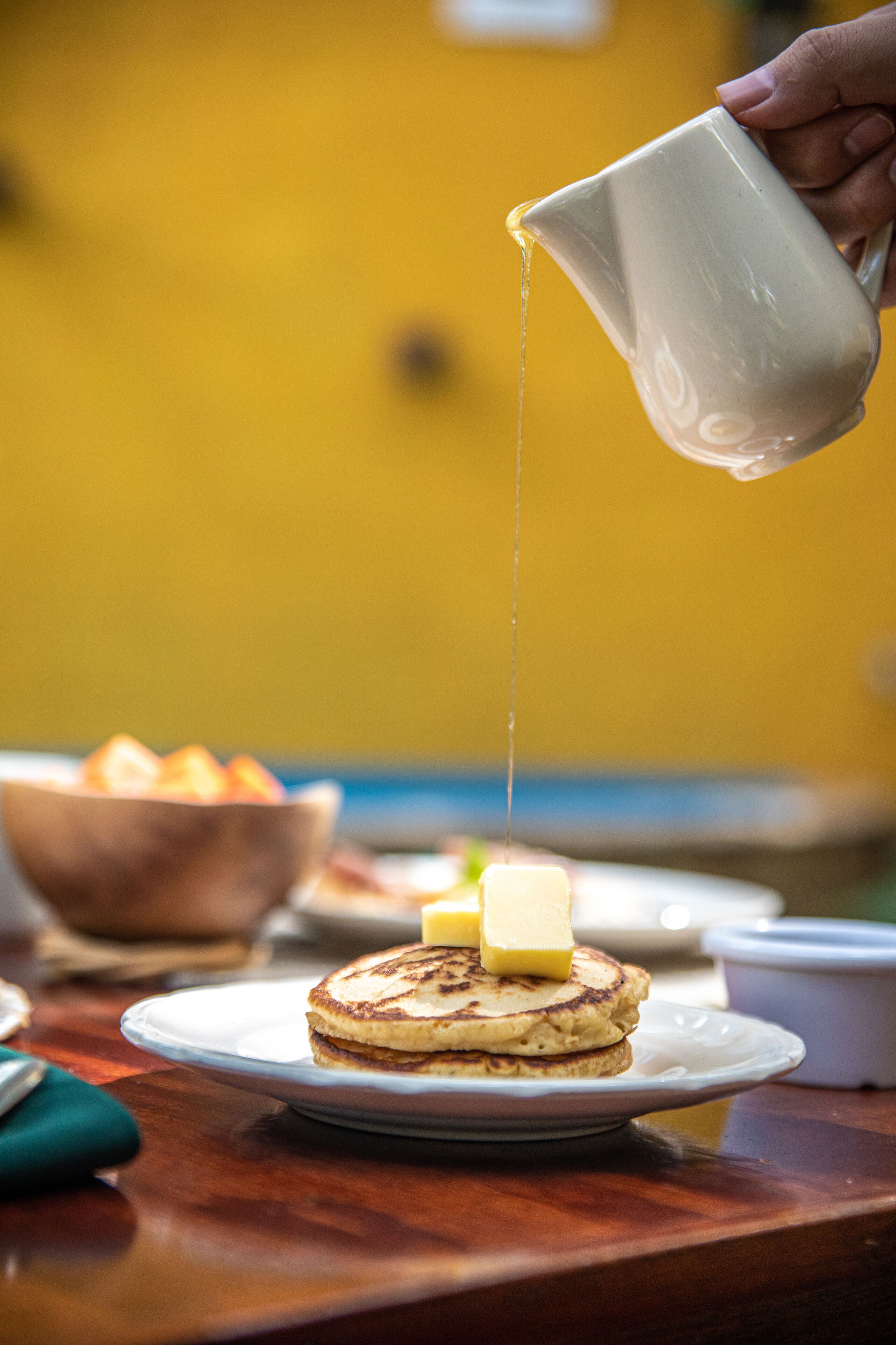 A hand pouring syrup onto a stack of pancakes on a plate.