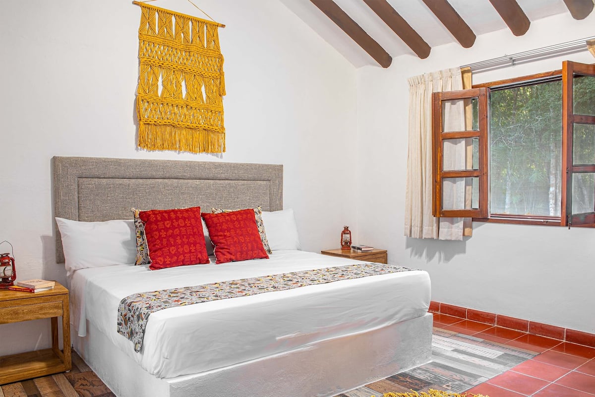A bedroom with a bed, a nightstand, a wall hanging, a window with curtains, and a wooden ceiling.