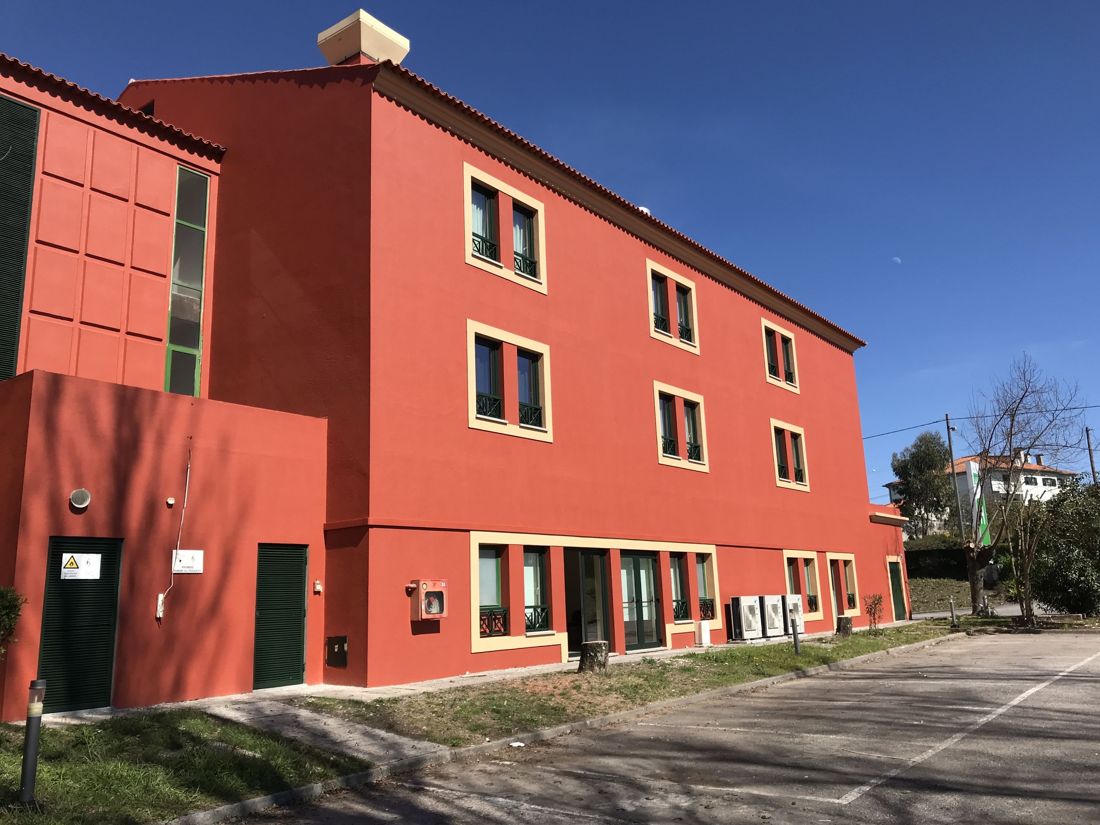 Un edificio rojo con varias ventanas y balcones, un estacionamiento al frente y un cielo azul despejado.