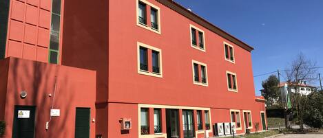 Un edificio rosso con numerose finestre e balconi, un parcheggio antistante e un cielo azzurro limpido.
