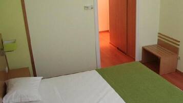 A single bed with a green blanket, a wooden bench, and a door leading to another room.